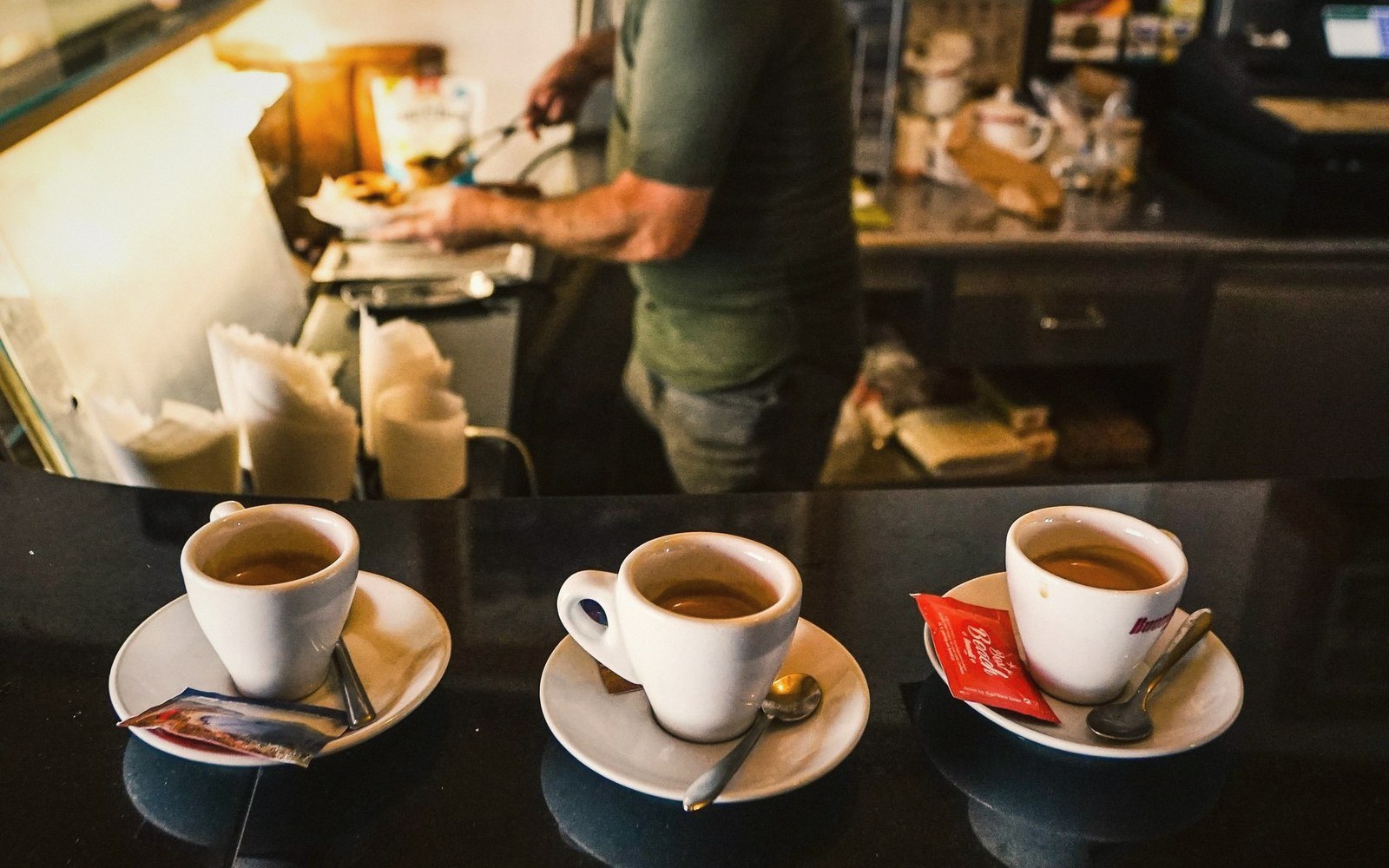 Three espresso cups on saucers lined up on a bar counter as a barista works behind it