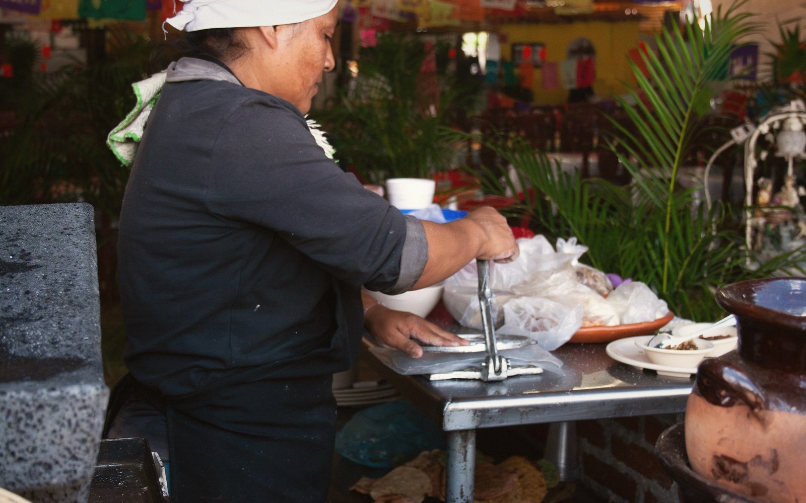 A woman uses a metal tortilla press to flatten corn dough at an outdoor stall