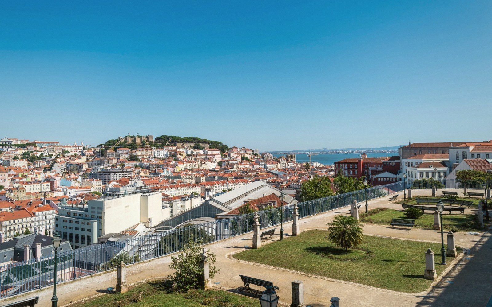 Terraced viewpoint garden overlooking Lisbon's rooftops with a hilltop castle and river beyond