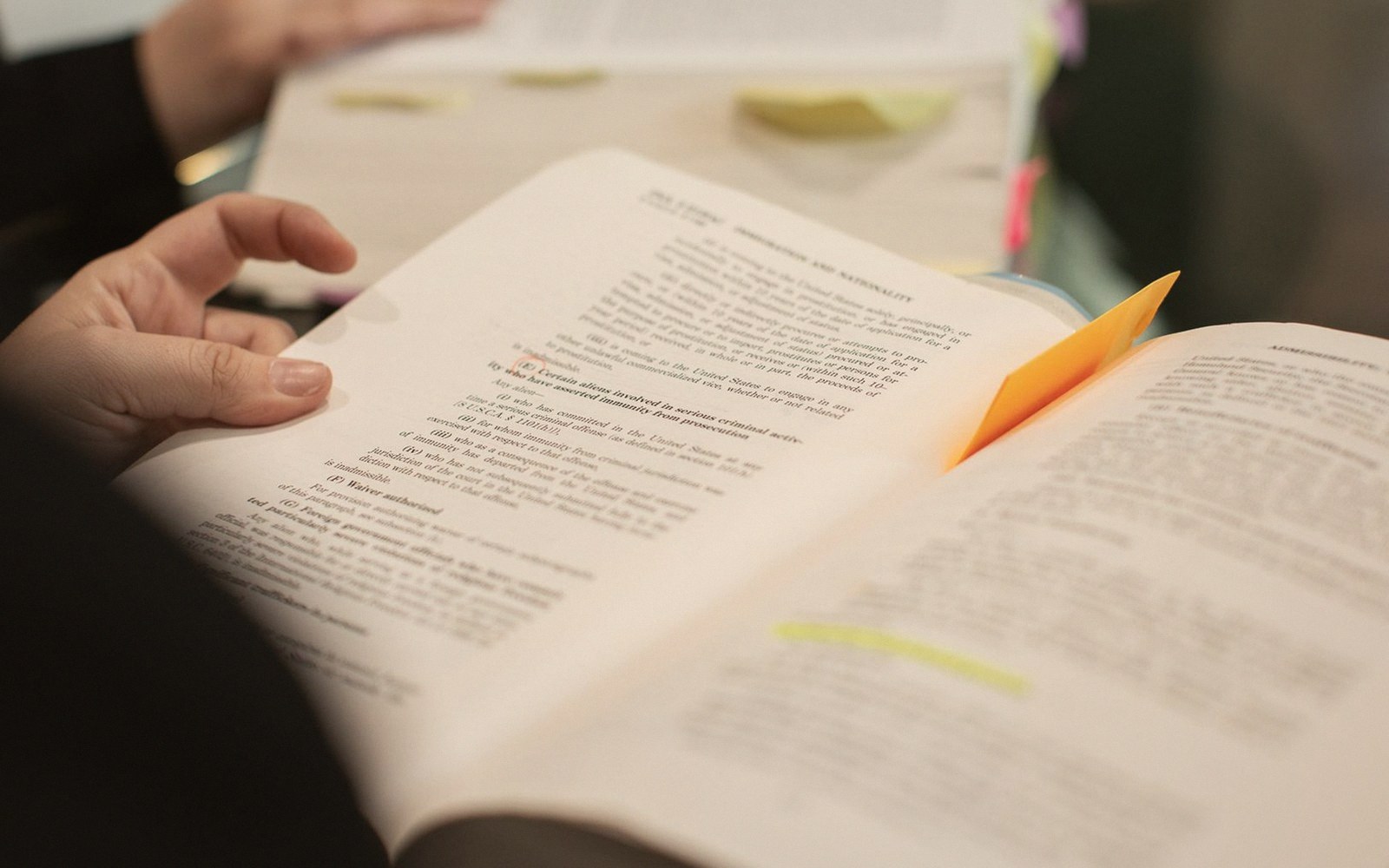 A hand holds open a legal textbook with an orange sticky note marking a page