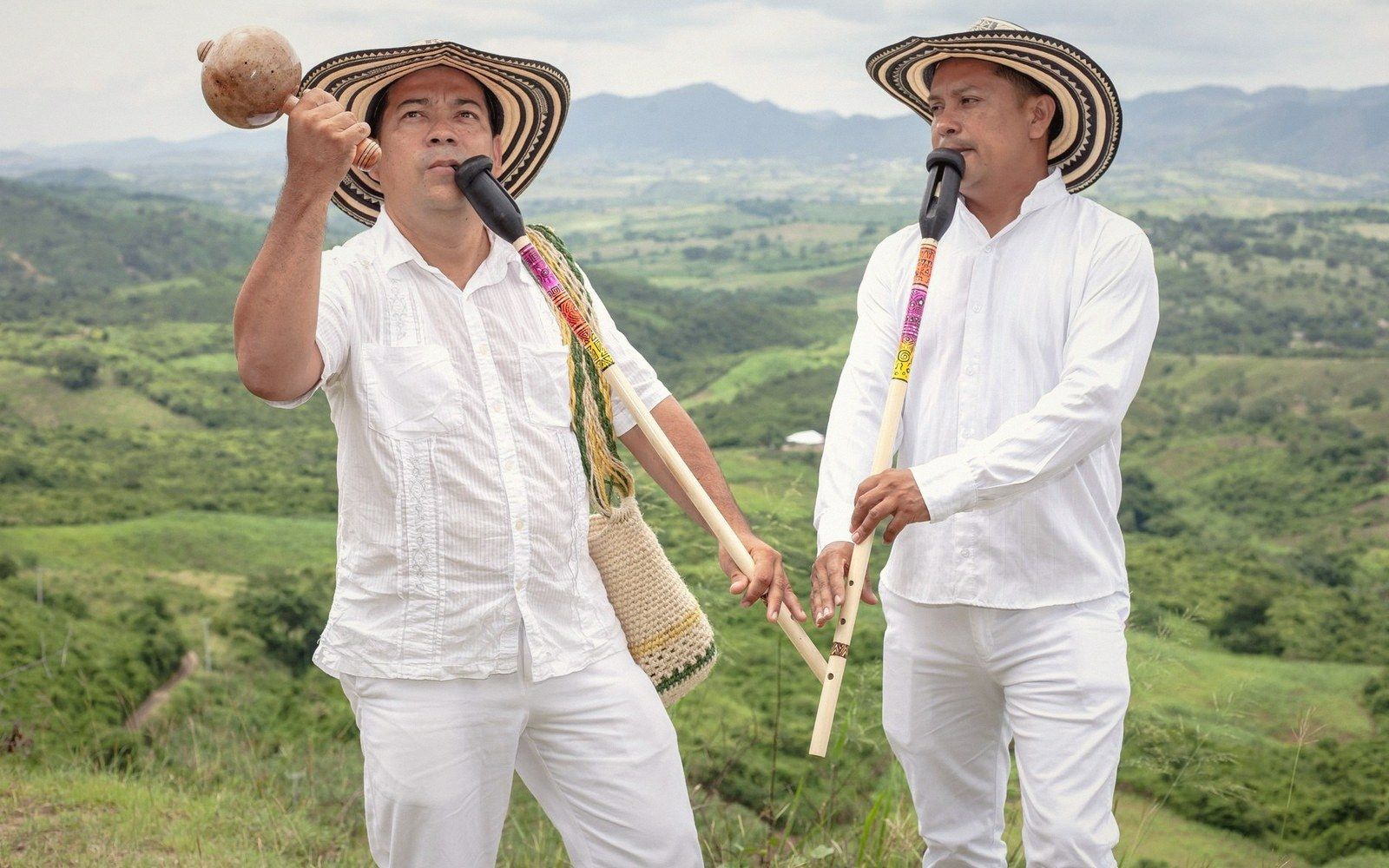 Two men in white shirts and sombreros play gaita flutes overlooking green hills